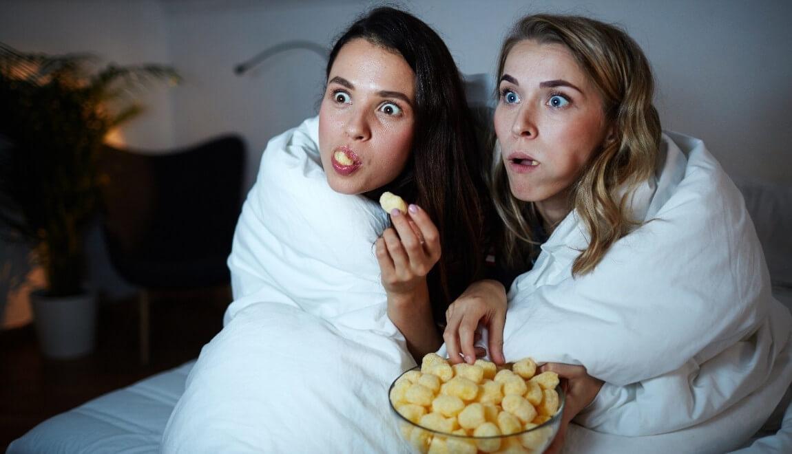 Two women wrapped in a blanket eating cheese puffs while watching a movie.