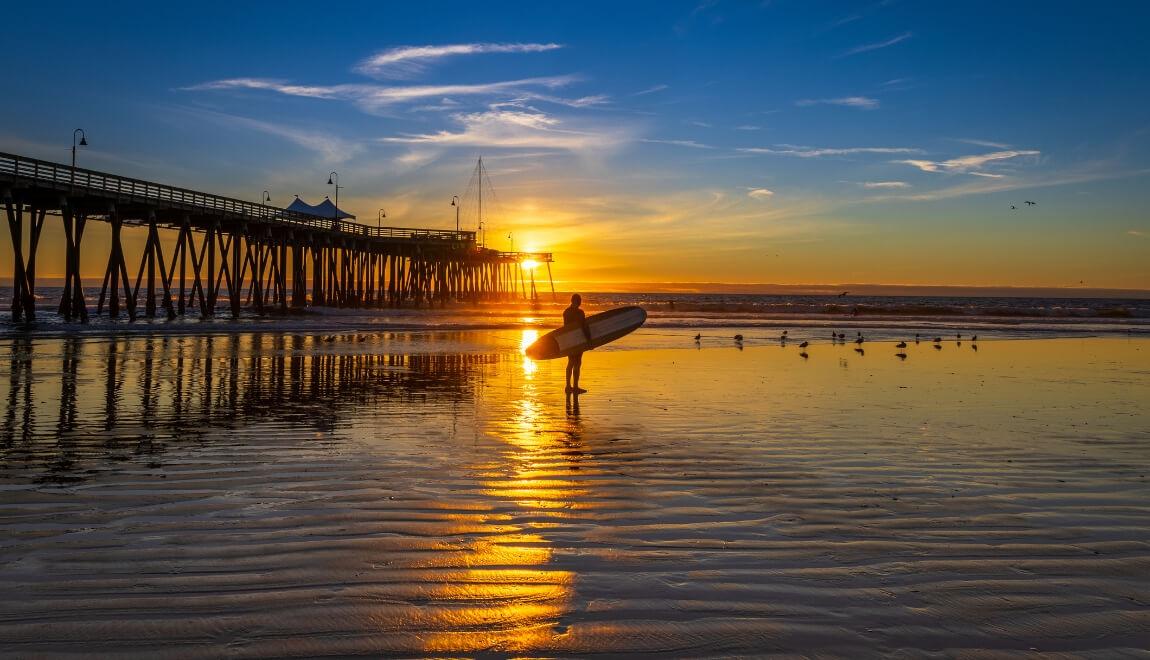 A surfer stands on Pismo Beach, California as the sun sets.