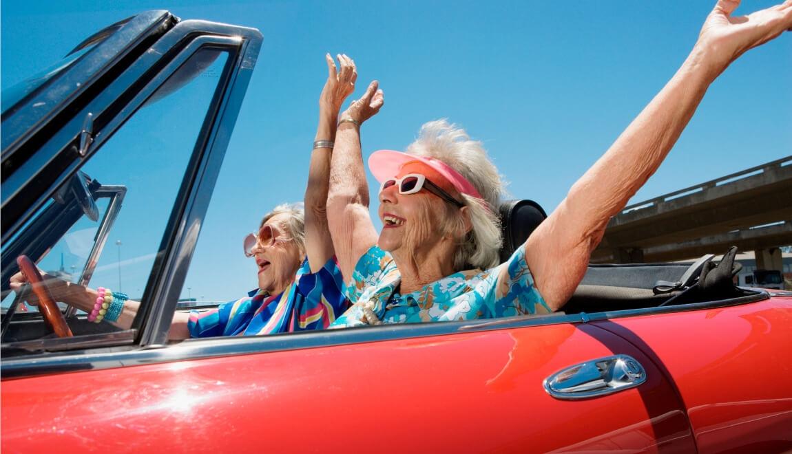 Two older women in a red convertible with their hands in the air.