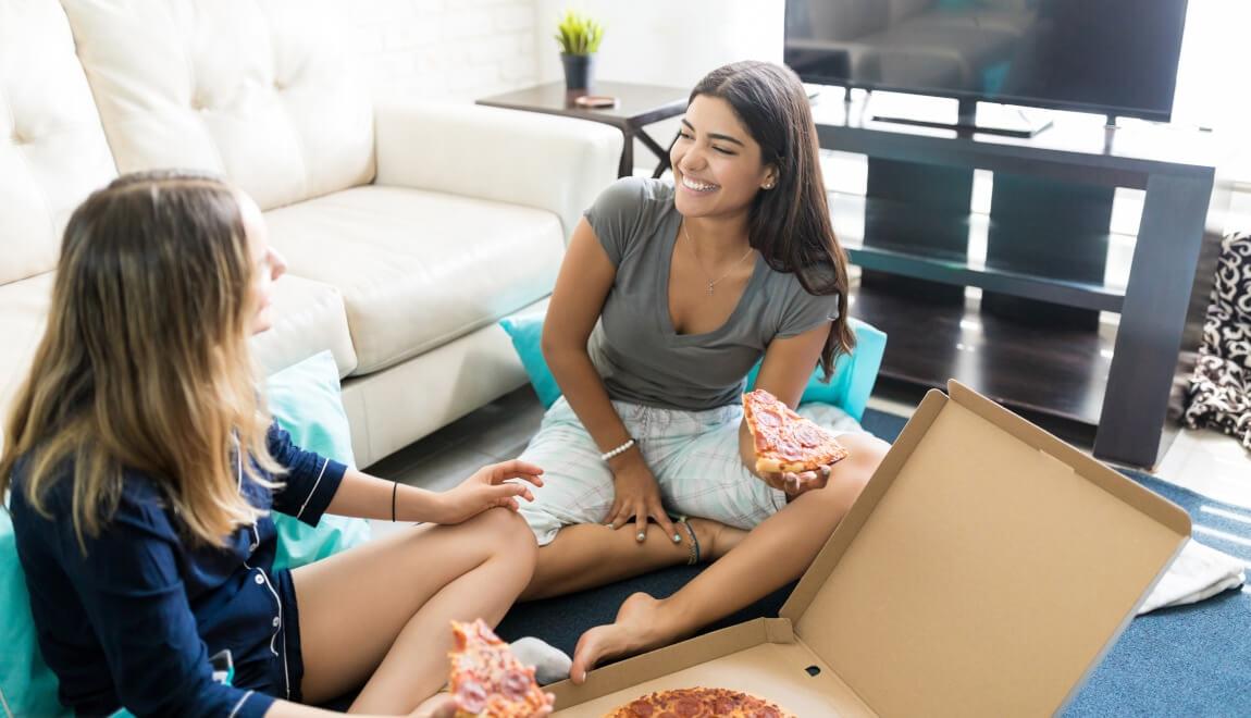 Two women having a conversation while sitting on the floor of a living room and eatting pizza.