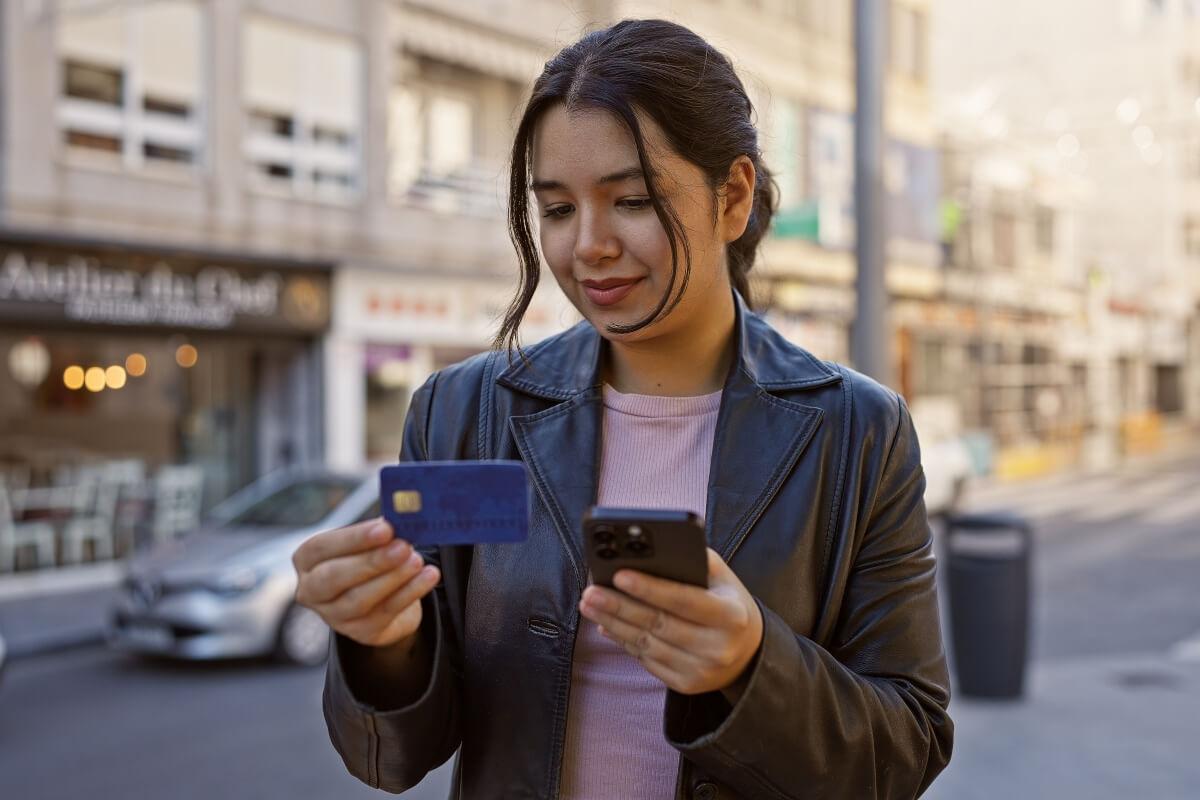 A young woman looks at her credit card on a sidewalk in an urban area.
