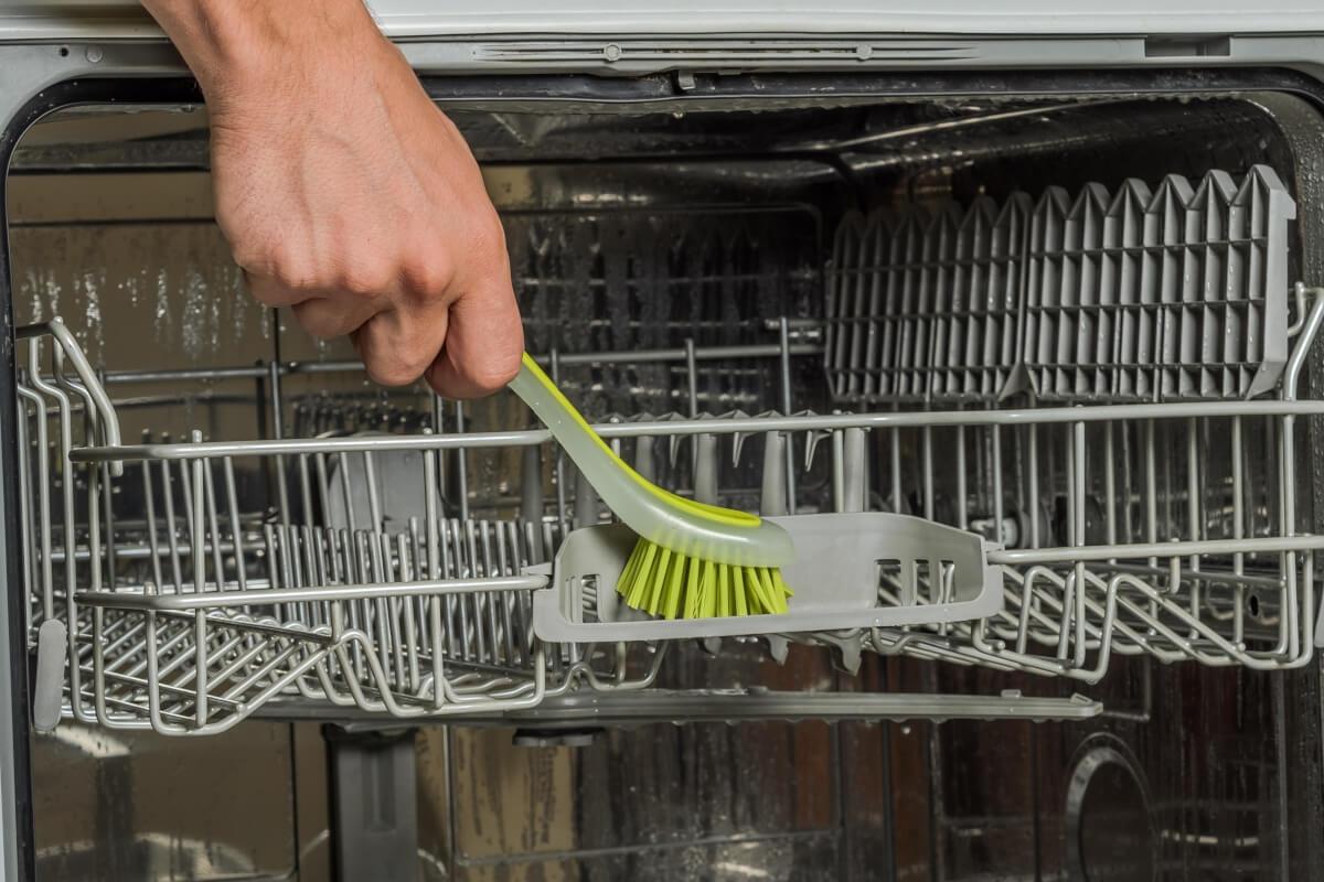 A person cleans the top rack of a dishwasher with a small scrub brush.