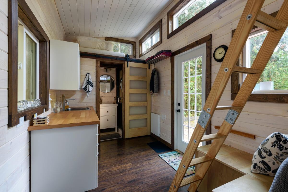 The inside of a tiny home shows a ladder in the foreground and a kitchenette and bathroom in the background.