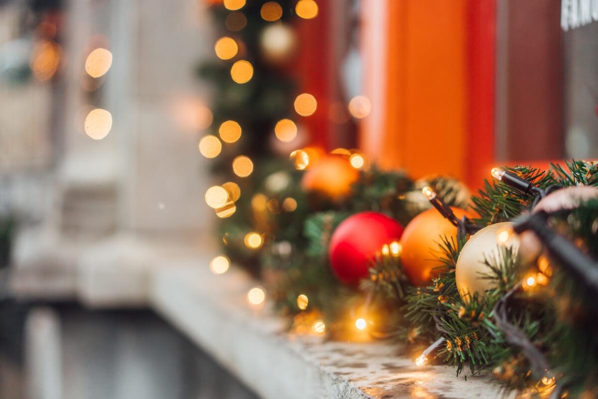 A close-up of a fireplace mantel shows an evergreen garland wrapped with lights and decorated with red, gold, and orange ornaments.