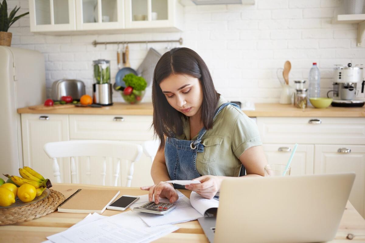 A young woman sits at a kitchen table making calculations.