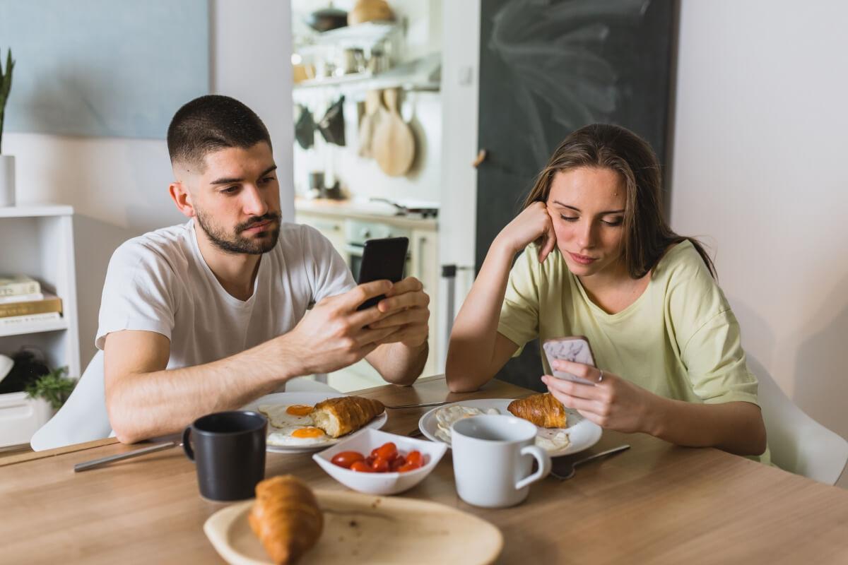 A young couple sits at a table eating breakfast while scrolling on their smartphones. 