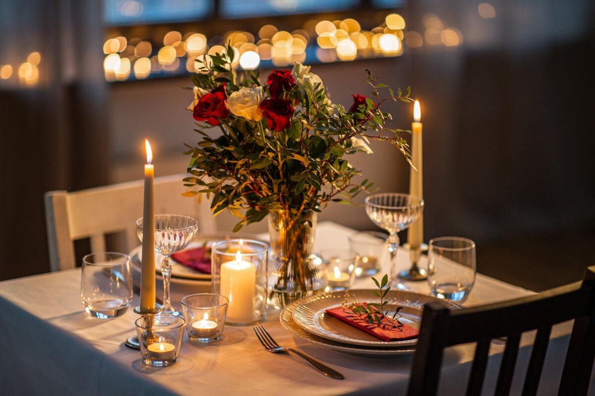 A table is set for two with a white tablecloth, candles, and a rose bouquet as a centerpiece.