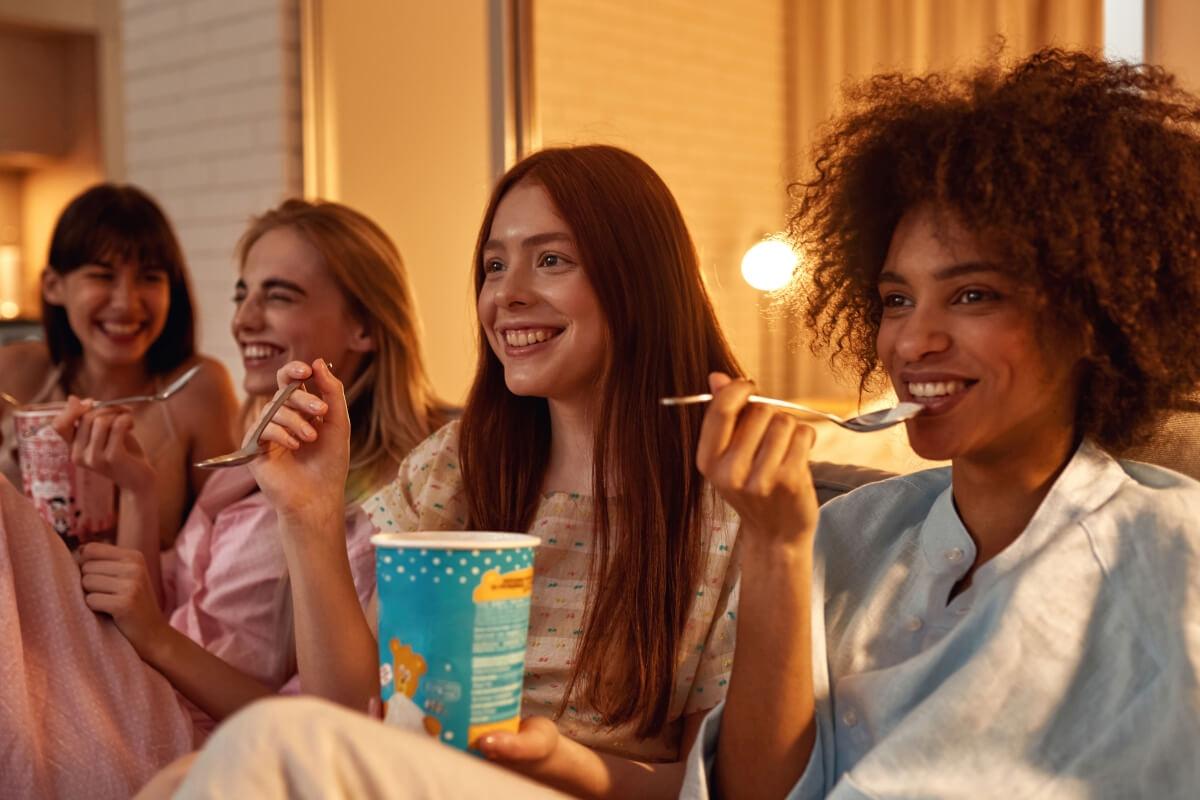 Four young women sit on a couch eating ice cream while watching something on a TV out of the frame.