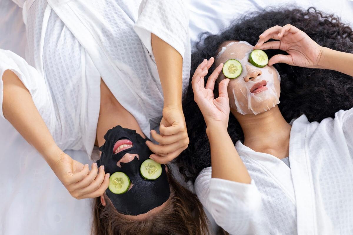 Two women lay on a bed with face masks on their skin and cucumber slices on their eyes.