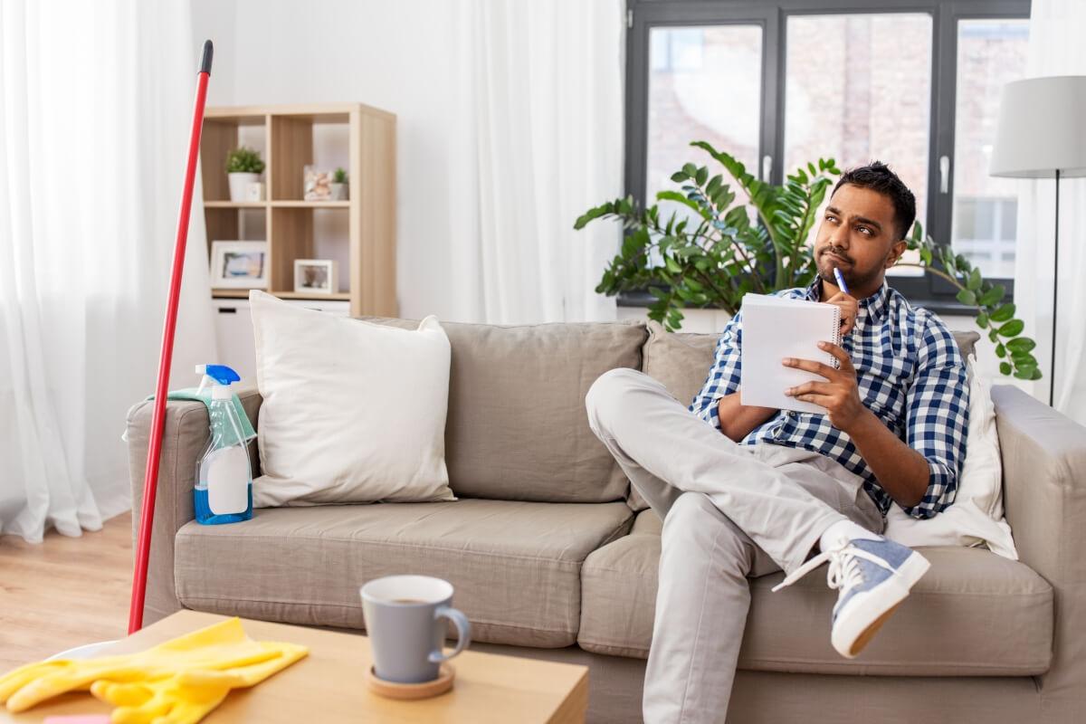 Man sitting on couch next to cleaning supplies as he comes up with cleaning to-do list.