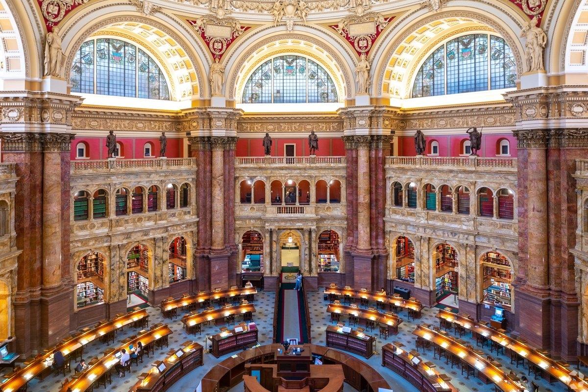 Interior view of an ornate reading room with marble columns, arched stained glass windows, statues, and curved rows of reading desks illuminated by warm lights.
