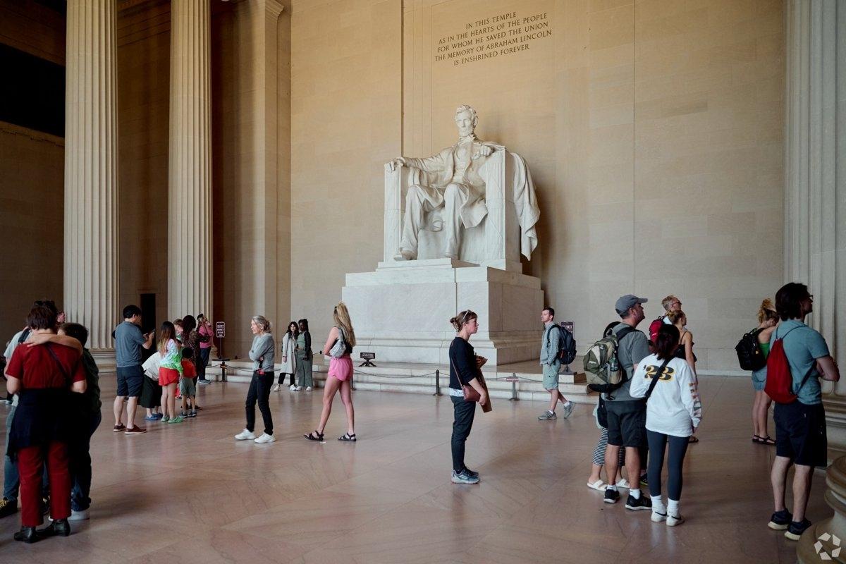 A large white marble statue of Abraham Lincoln seated on a throne inside a grand pillared hall, with the inscription “IN THIS TEMPLE…THE MEMORY OF ABRAHAM LINCOLN IS ENSHRINED FOREVER” above and visitors standing nearby.