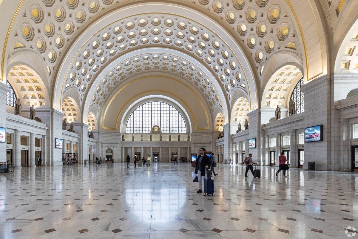 A spacious grand hall with high vaulted ceilings featuring coffered arches, statues along the walls, and travelers pulling rolling suitcases.