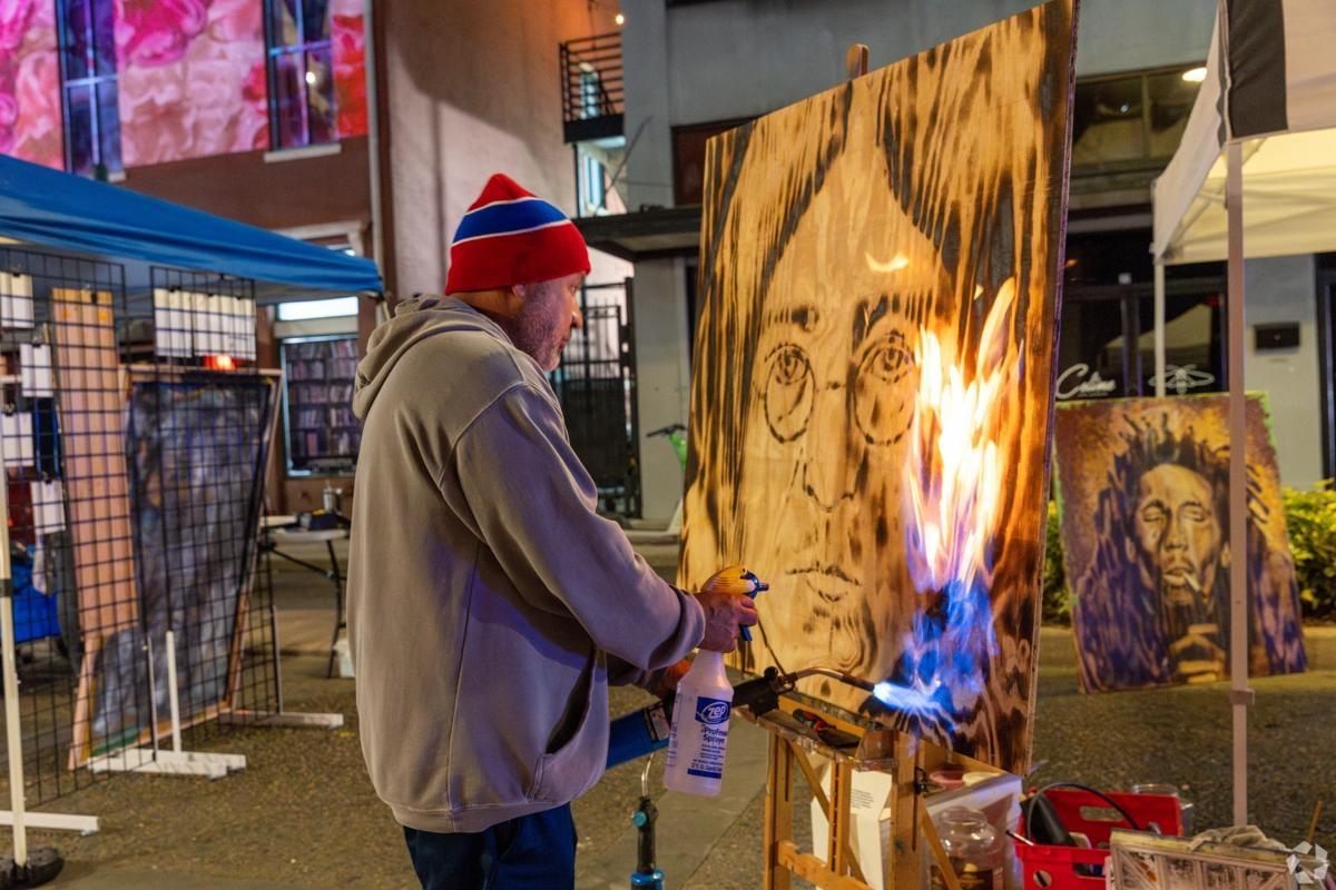 Local street artist using fire to create a wood-burned portrait at an urban art fair, highlighting vibrant culture near downtown apartment communities.