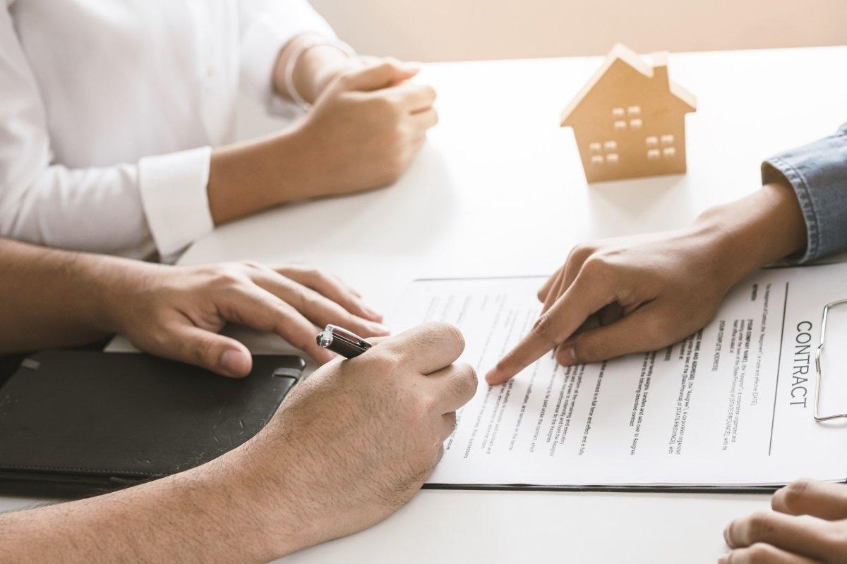 Person signing contract while another person points at specific points and someone sits next to them.