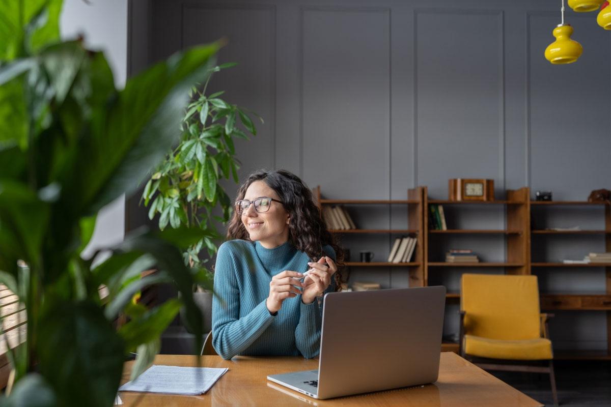 Young lady gazing out of apartment window as she sits in front of laptop.