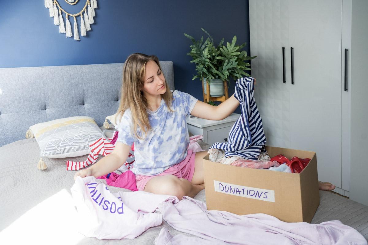 A young woman sits on her bed and considers which of her clothes to keep and to donate.