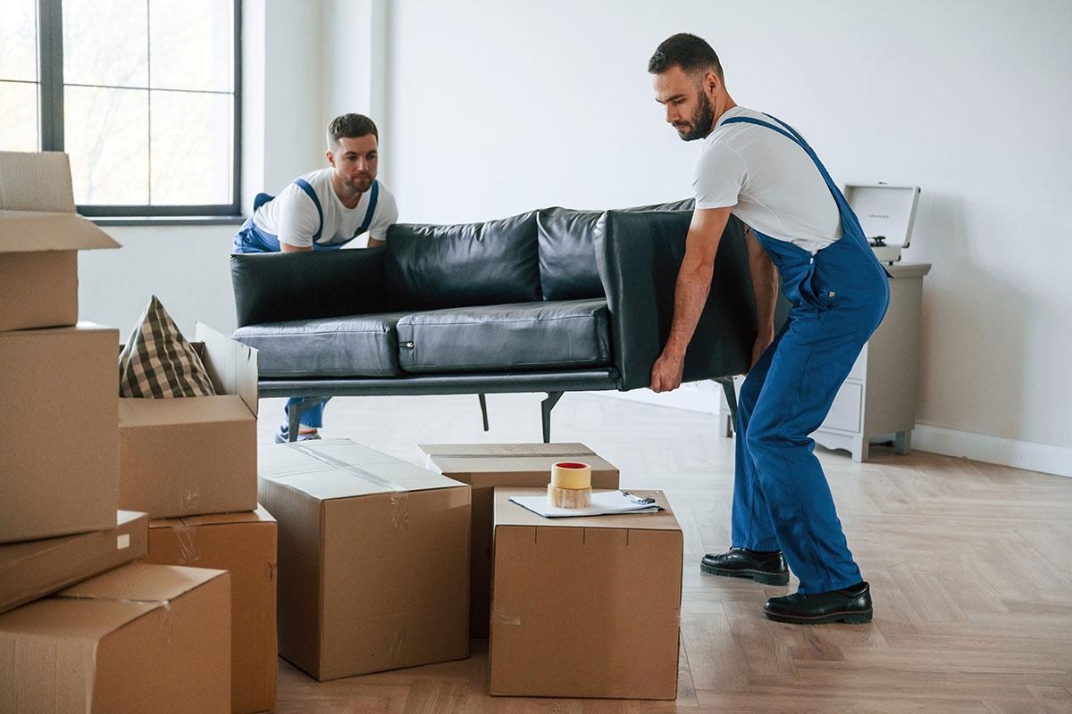 Movers carrying black leather couch out of apartment filled with boxes.