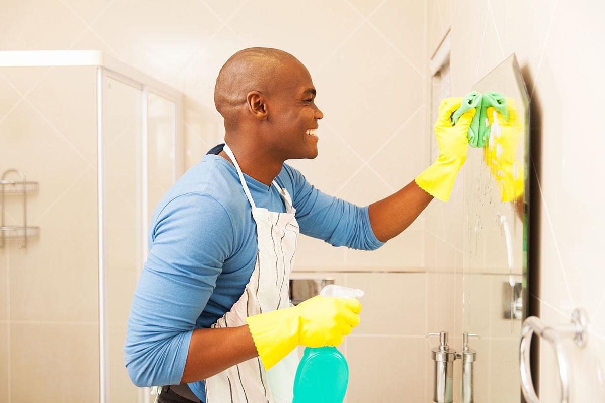 Man in cleaning gear as he sprays and cleans bathroom mirror.