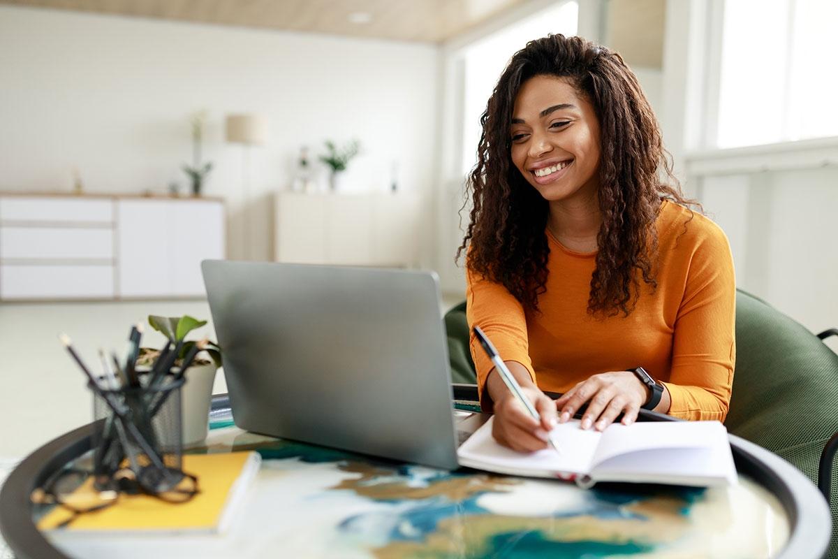 Woman taking notes as she uses her laptop and smiles.