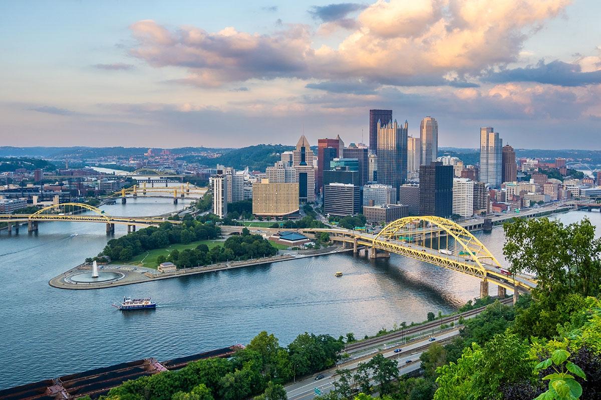 Downtown Pittsburgh sits where multiple rivers meet, highlighted by yellow bridges crossing the water and a skyline of tall buildings rising from the riverbanks.
