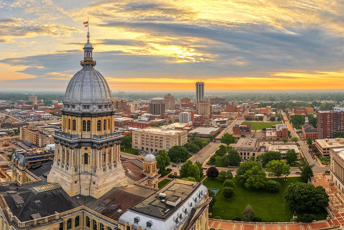 The Illinois State Capitol building in Springfield at sunset, with its large domed roof in the foreground and the surrounding downtown cityscape stretching into the distance under a colorful sky.