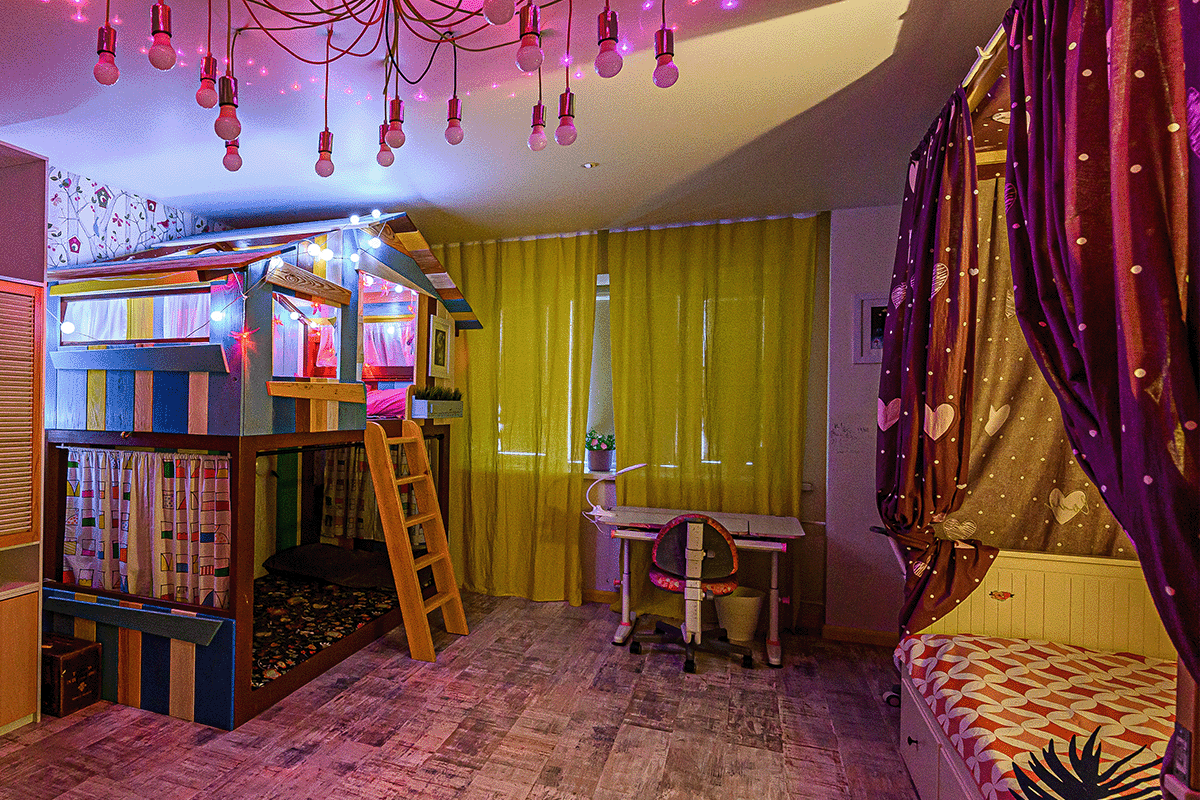 chidlren's bedroom with a patterned rug. There is a light fixture with many lightbulbs and purple hues. The curtains over the windows are yellow, while the curtains over the children's bed are black with heart patterns. To the left of the room, there is a constructed house simulating a treehouse. 