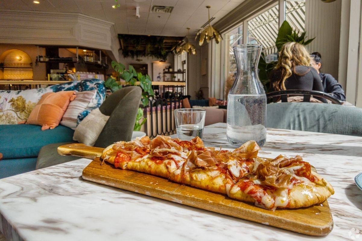 A wooden platter holding a long pizza topped with prosciutto