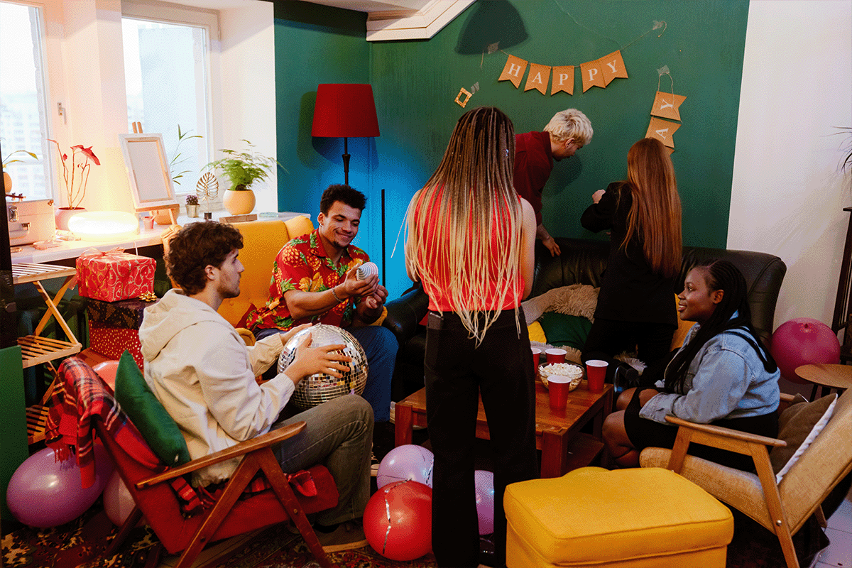 There are six people gathered in a dark green room. There are balloons across the floor and chairs that are red, black, beige, and red. The decor is ecclectic and very busy in the small room.