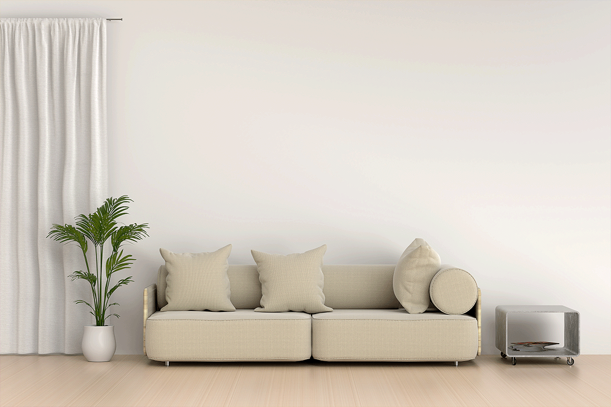 a neutral living room with a beige couch, bare walls, and a medium sized plant to its left.