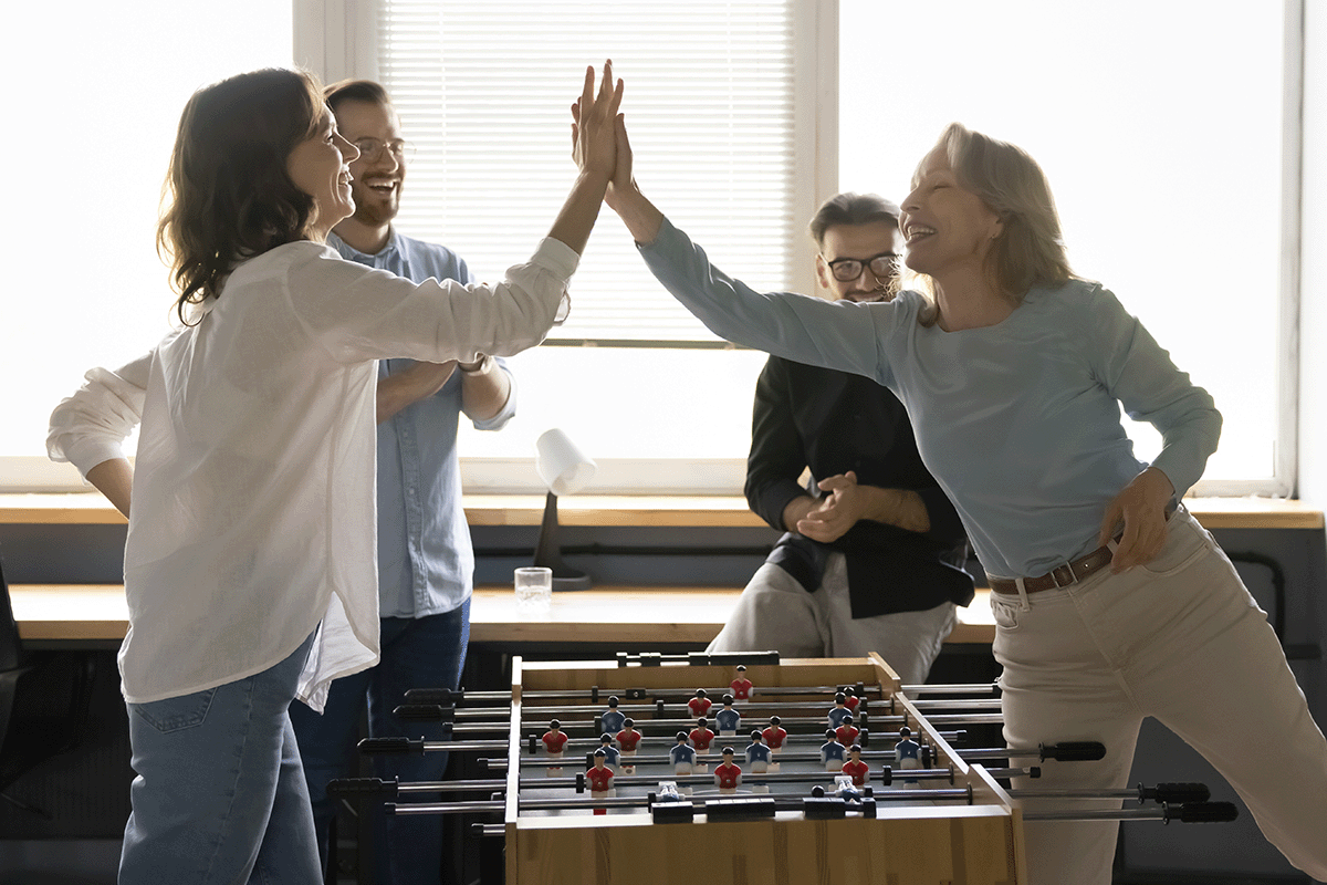 Two women high-five each other over a game of foosball. There are two men in the back clapping.