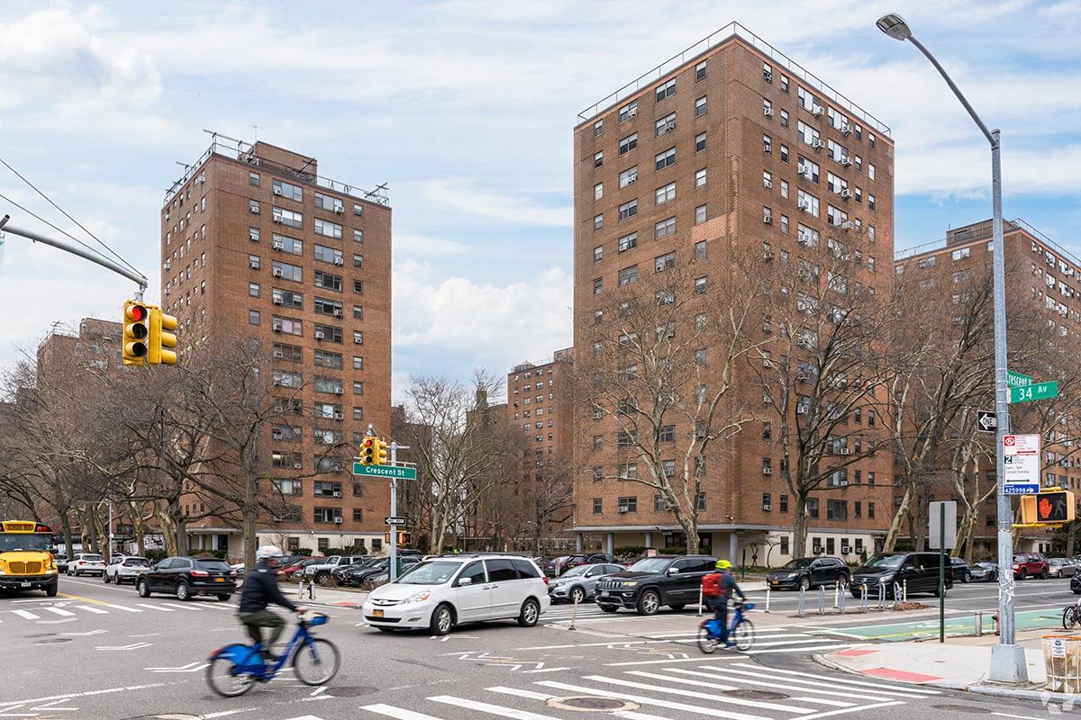 Two people riding Citi Bikes on a bike lane on a busy street, past apartments in Astoria.
