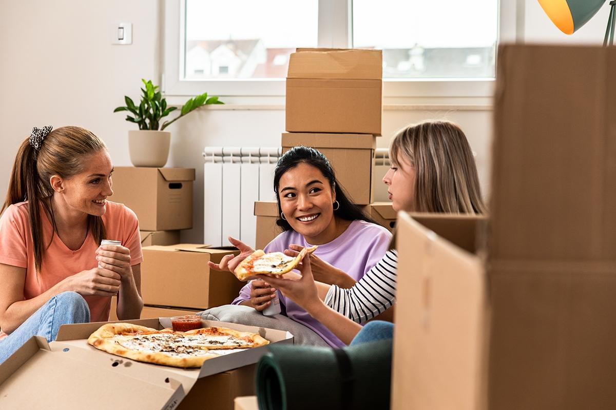 Three women sitting and eating pizza as they are surrounded by moving boxes.