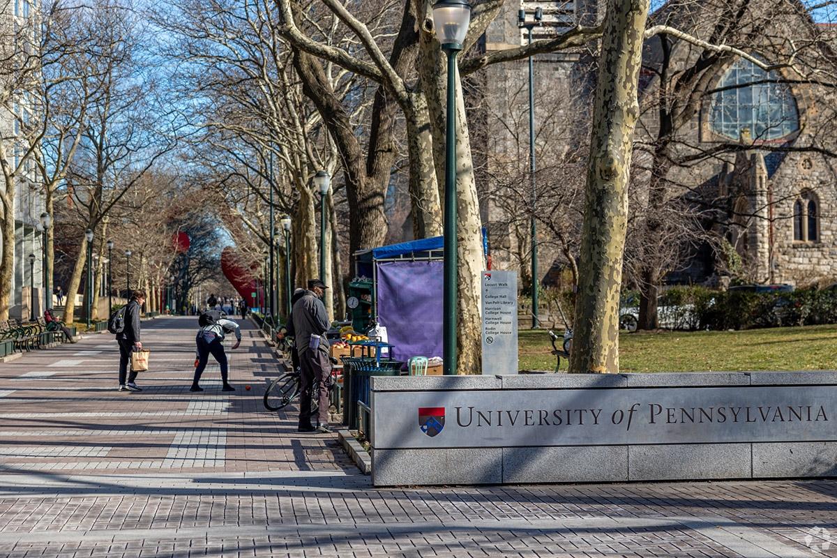 People next to a stone sign displaying University of Pennsylvania on a main campus thoroughfare.