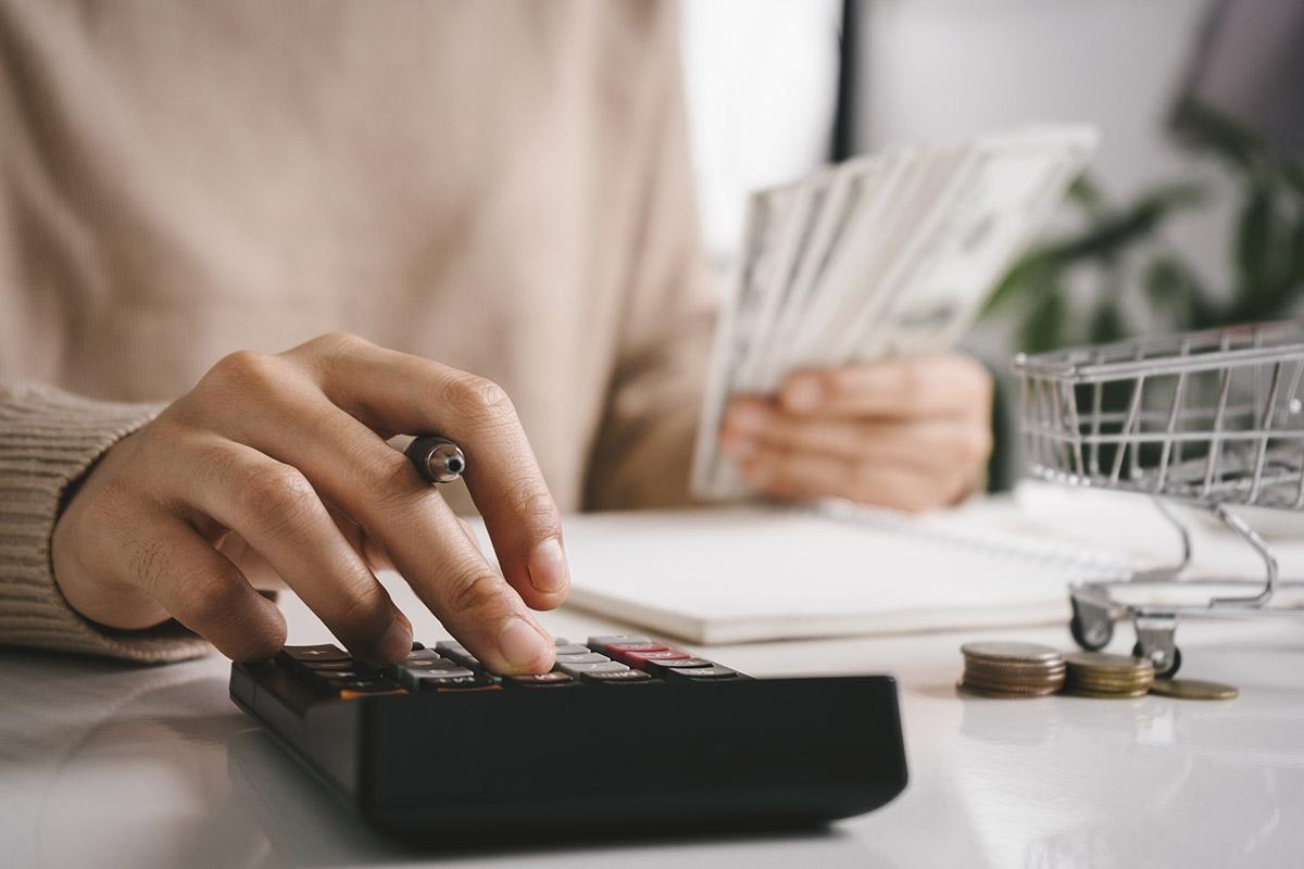 Hand holding pen and using calculator as the other hand holds money in the background.