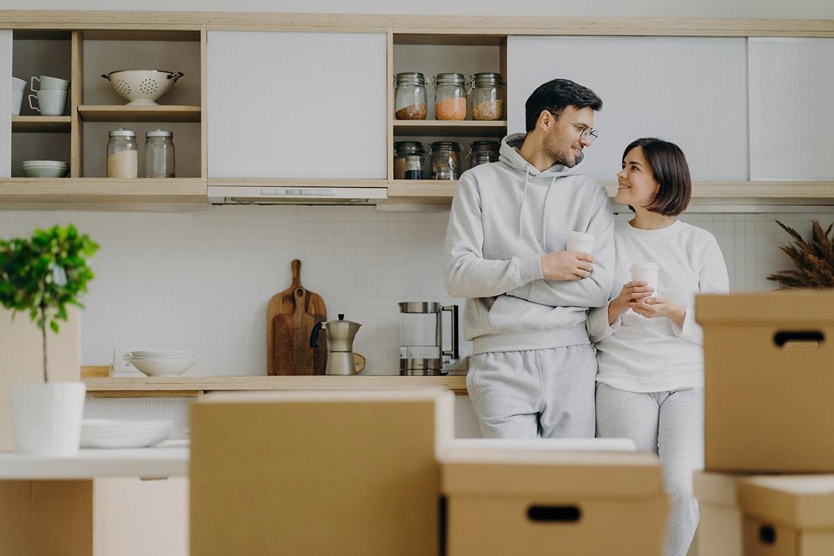 A man and a woman drinking coffee in their apartment's kitchen as moving boxes sit in front of them.