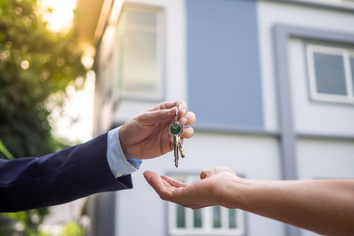 A hand handing over a set of keys to another hand with a house in the background.