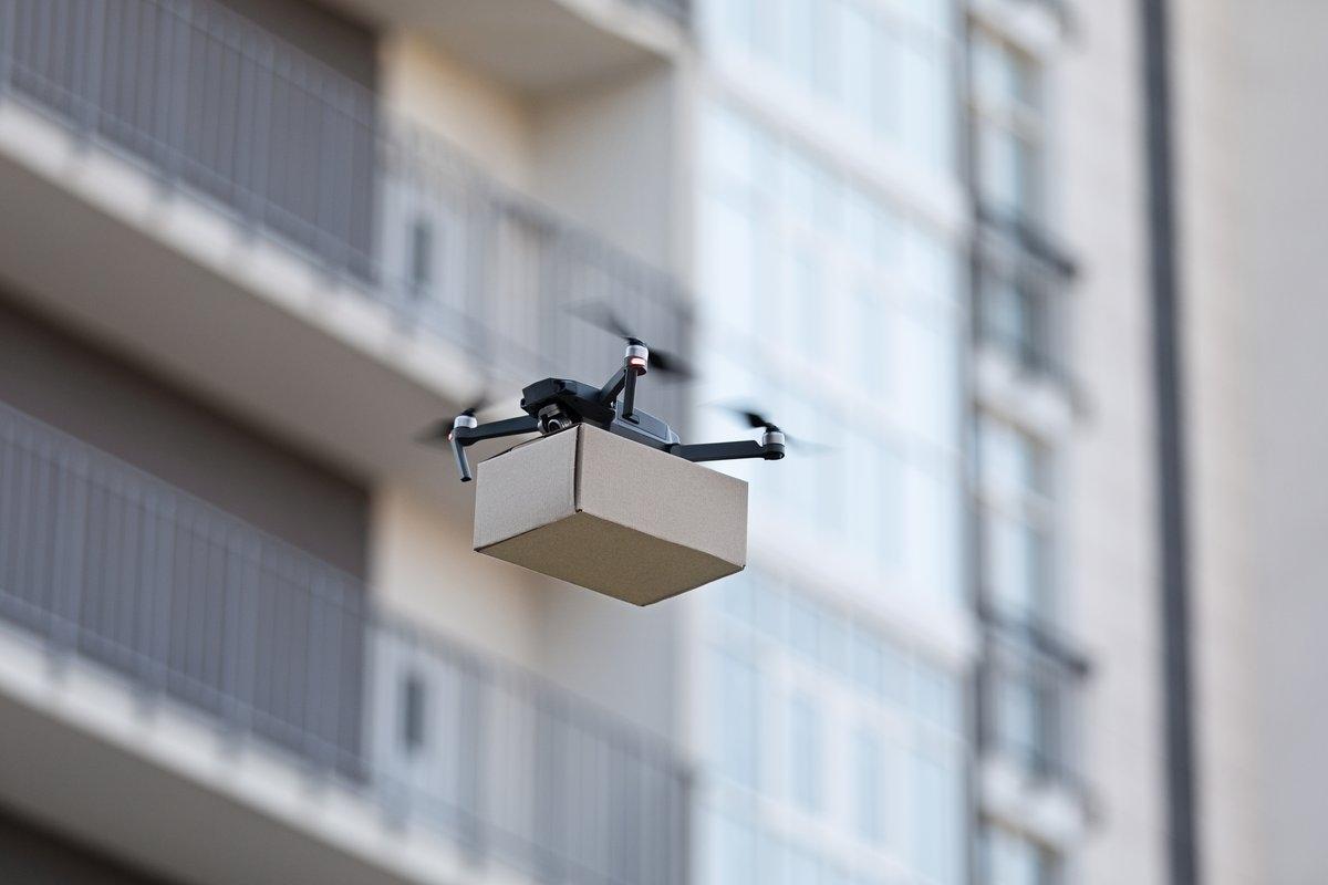 A drone holds a package in front of an apartment building.