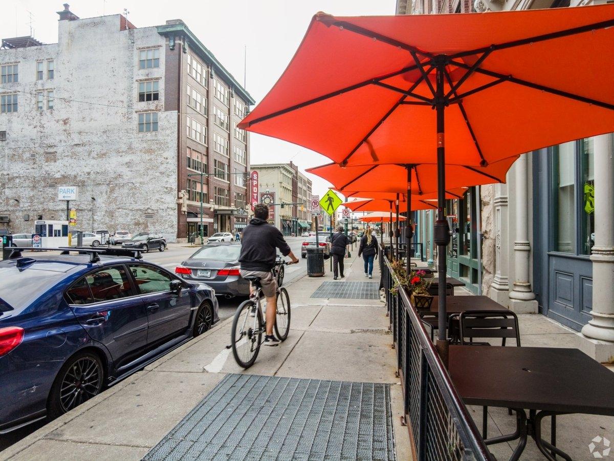 A person bikes on the sidewalk, past the outdoor tables under umbrellas of a restaurant.