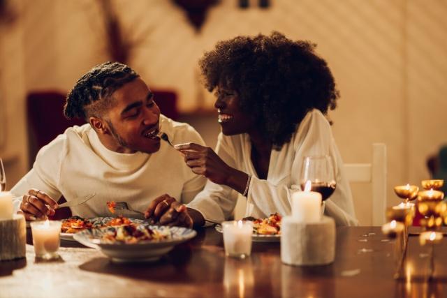 A smiling young woman gives her partner a bite of her dinner.