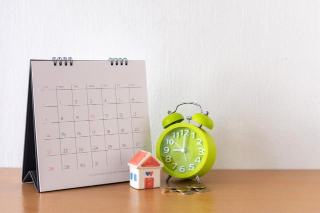 Coins sit in front of a lime green alarm clock that is next to a calendar that has a small house in front of it.