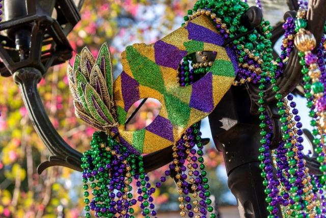 A classic Mardi Gras mask hangs on a lamppost alongside beaded necklaces in purple, green, and gold.