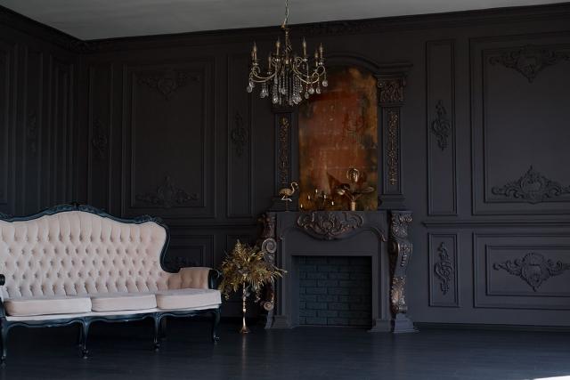 An opulent living room showcases paneled walls, a chandelier, a carved fireplace that are all black as well as a pale upholstered sofa in low light.