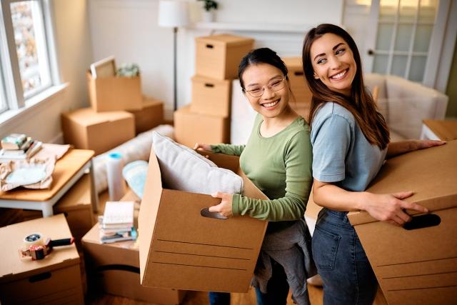 Two women smiling and standing back to back while holding boxes as they move into their new apartment.