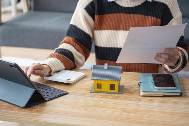 Woman typing on laptop while holding papers with a calculator, model house, phone, and notebook in front of her.