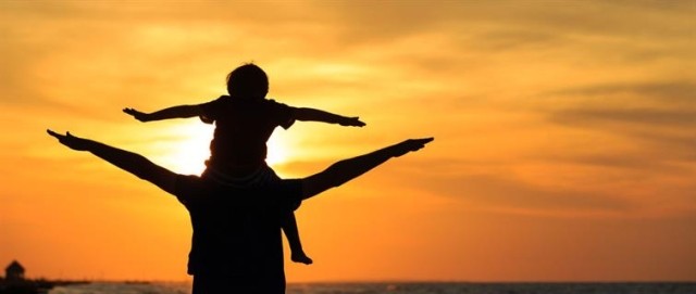 A child sits on a parent's shoulders, arms outstretched as they face the sunset.