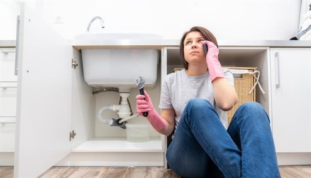 A woman sitting on the floor next to her sink calling maintenance.
