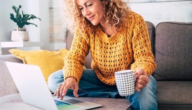 A woman in a yellow sweater holds a coffee cup while browsing on a laptop computer.