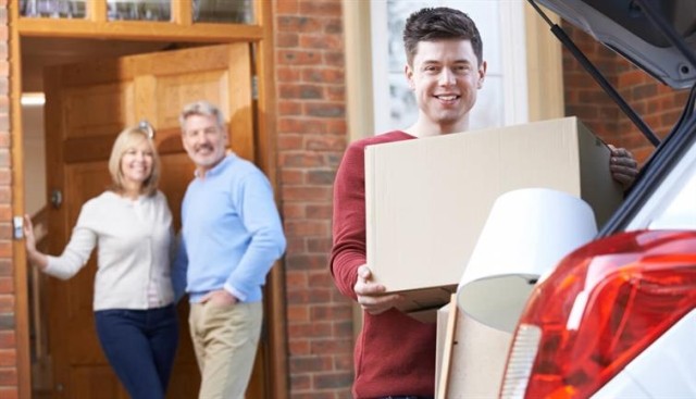 Adult child loads car with boxes as his parents watch from the doorway.
