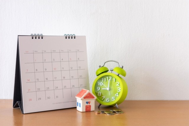 Coins sit in front of a lime green alarm clock that is next to a calendar that has a small house in front of it.